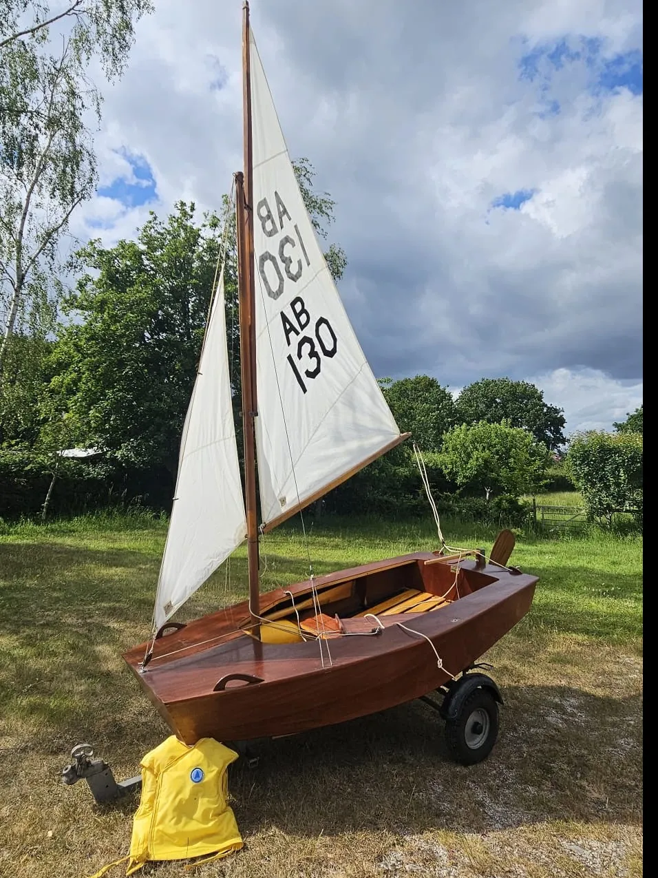 An original Poole AB rigged on her trailer in a field, varnished mahogany hull, Terylene sails on wooden spars, and a yellow lifejacket propped against the trailer hitch.
