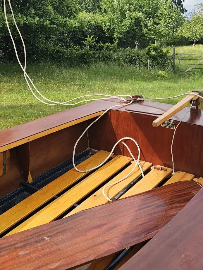 Looking into the barn find's cockpit — original slatted floorboards, varnished side benches, halyard coiled across the deck, and the builder's plaque still fixed to the bulkhead.