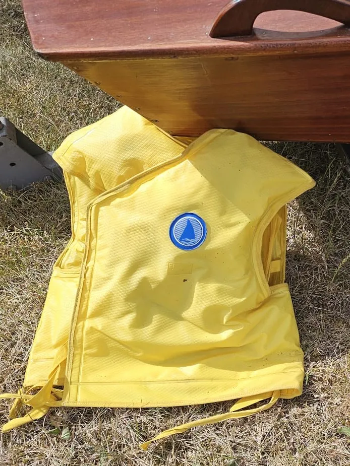 A yellow 1960s lifejacket with its ties, lying on the grass beside the bow of a varnished mahogany hull.
