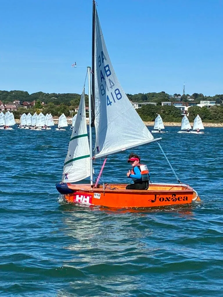 FoxSea, an AB with an orange hull and her name in black script along the side, sailing in Poole Harbour with a young helm giving a thumbs up and a fleet of dinghies behind.