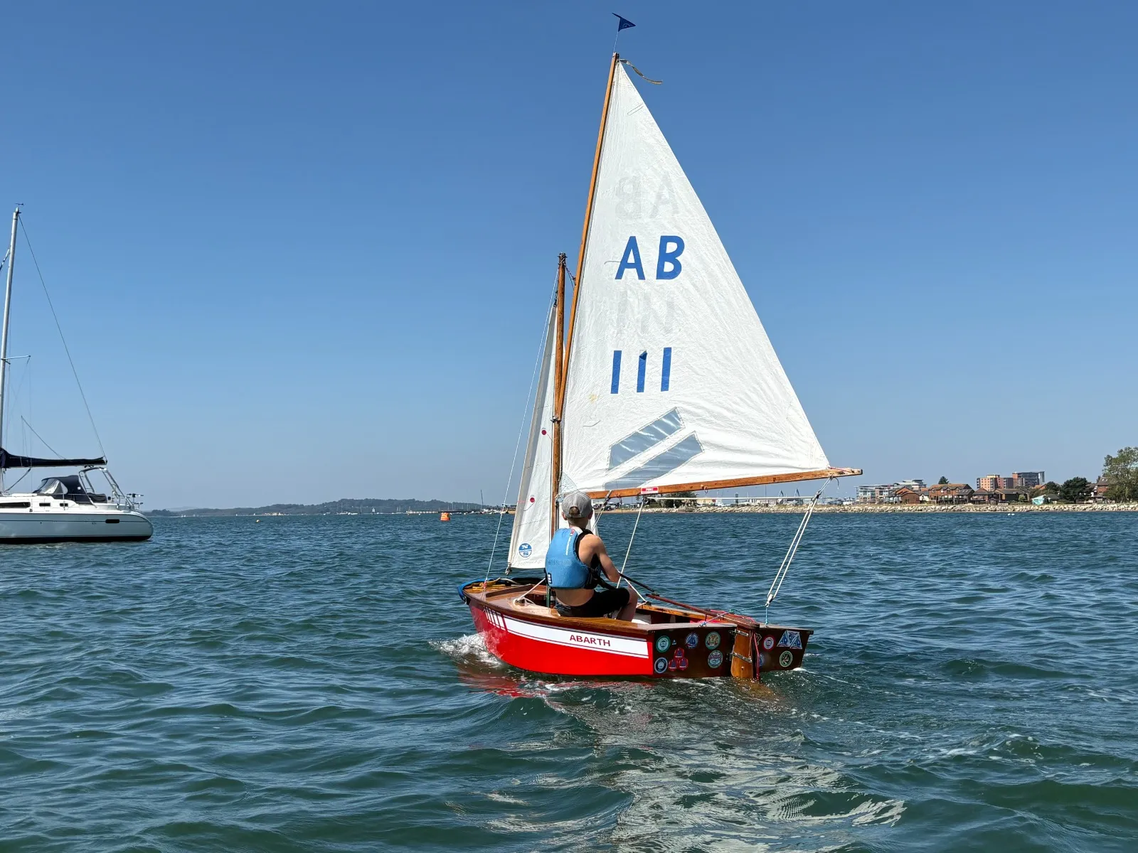 Abarth, a red-hulled AB, sailing away across the harbour, mainsail numbered 111 and stickers along her transom, one young sailor aboard.