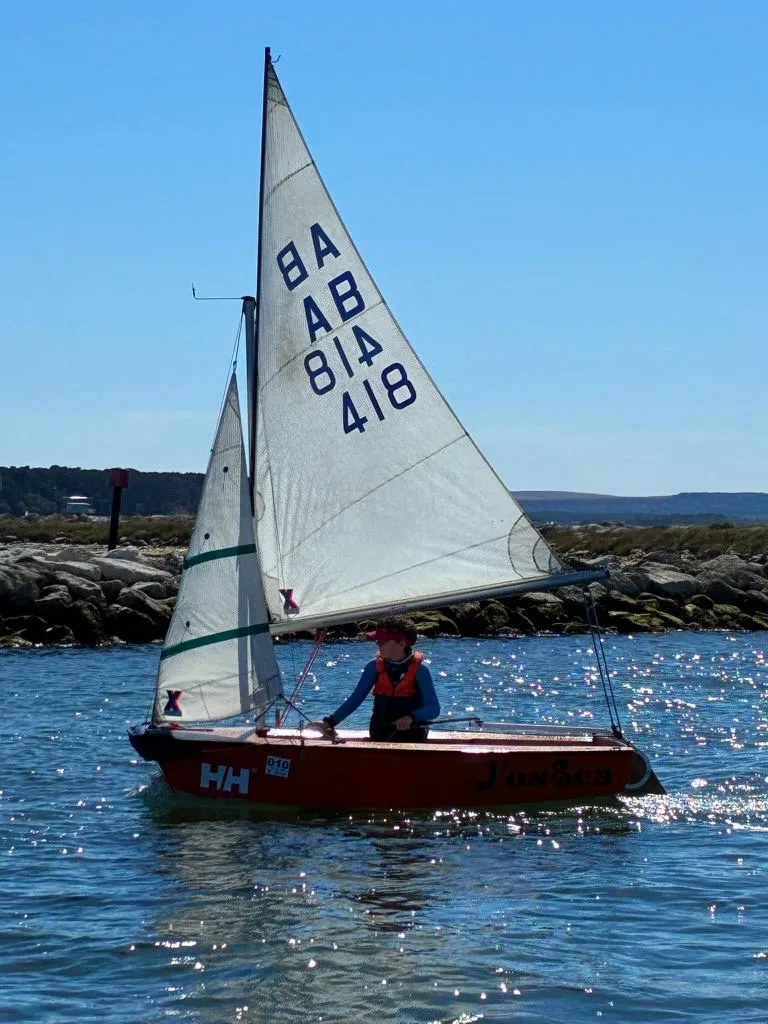 FoxSea sailing past a rock breakwater in bright sun, mainsail numbered 418 and a green-striped jib, one young sailor aboard.
