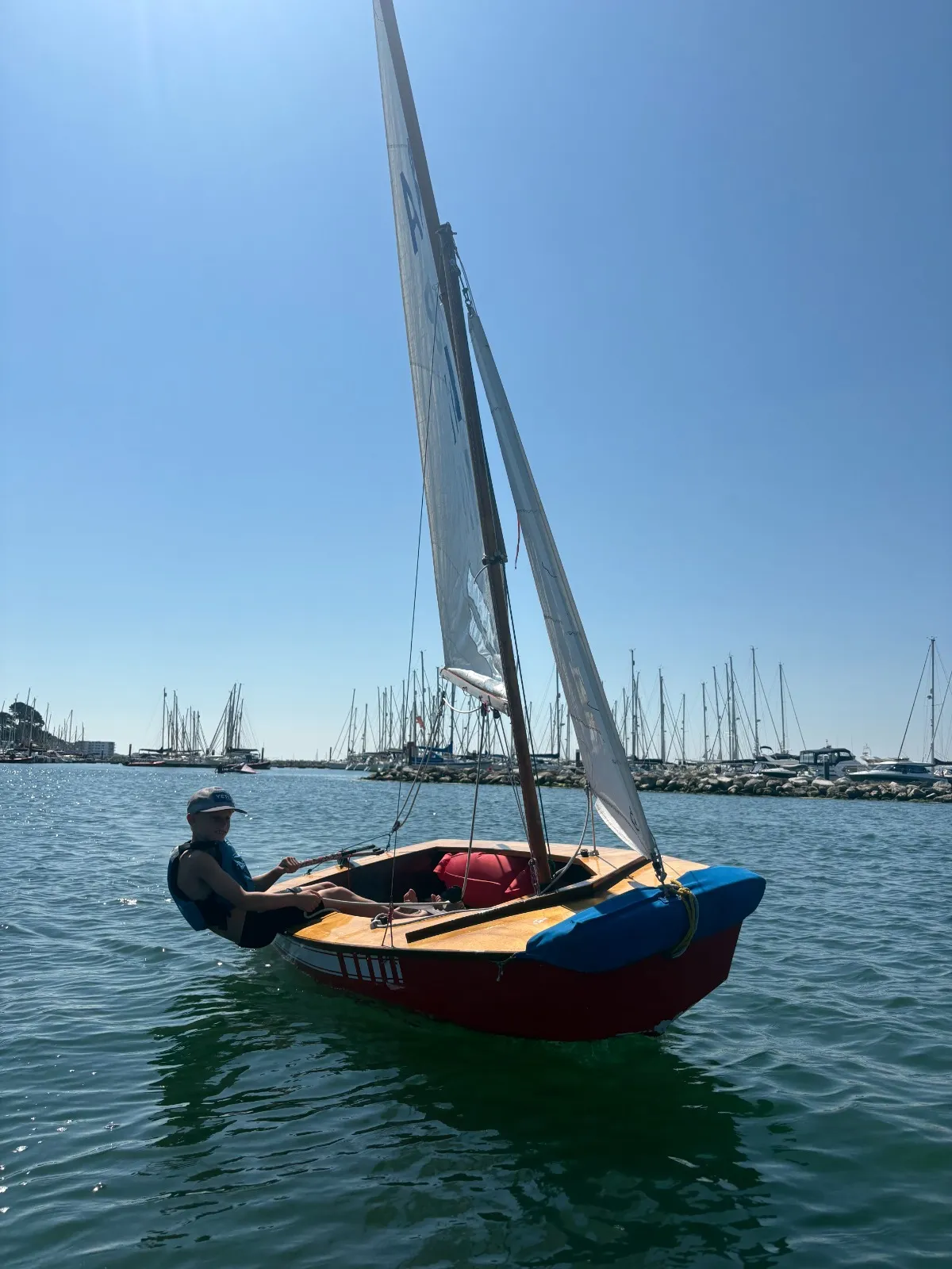 Abarth heeling gently as she sails off the marina, a young sailor at the tiller and a blue cover over the foredeck.