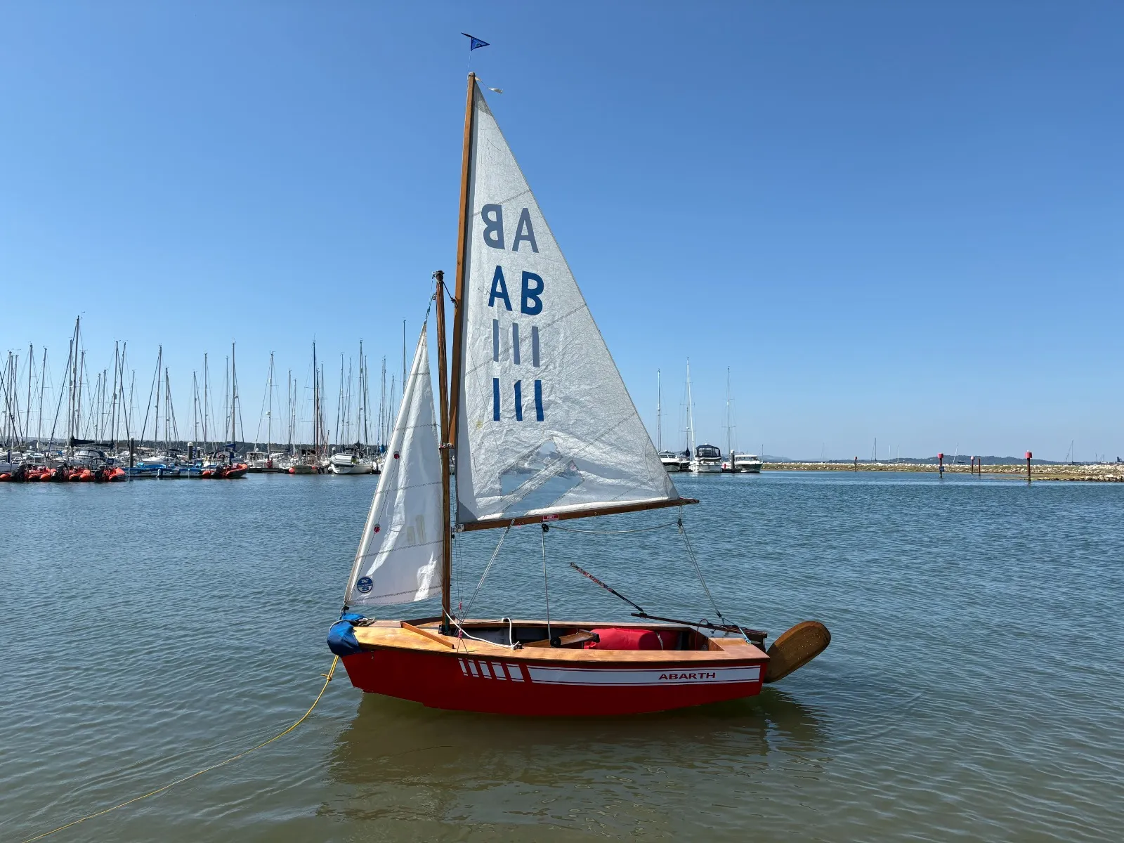 Abarth afloat in calm water off a marina, sails up, red topsides with a white stripe.