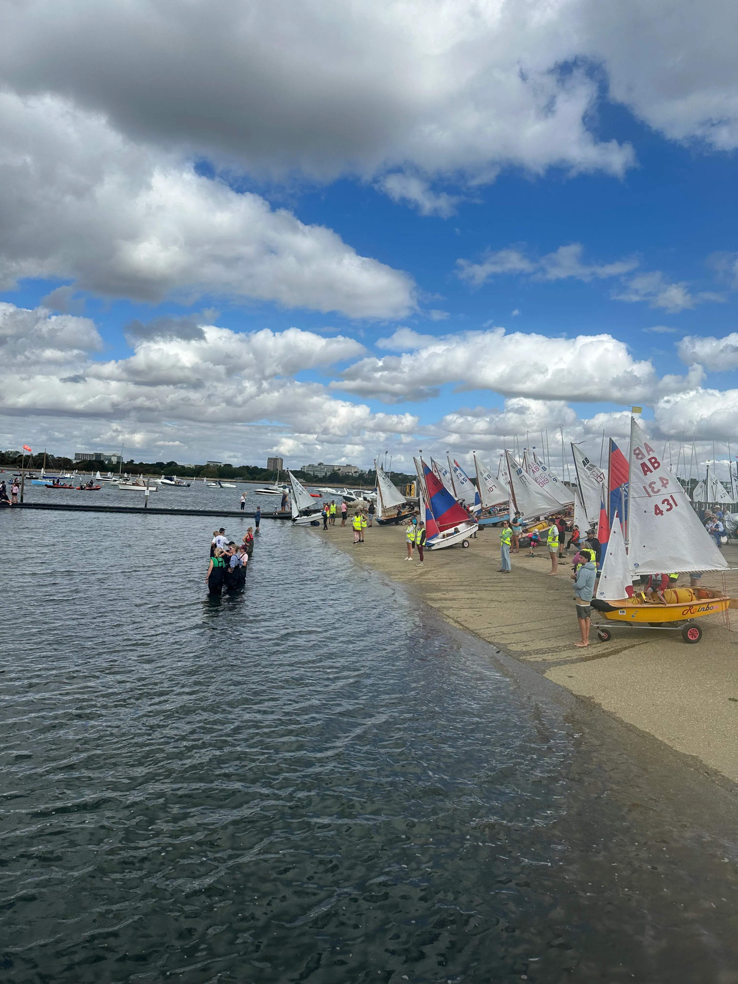 Children in wetsuits and buoyancy aids launching an AB off the slipway at Parkstone, with the rest of the fleet rigged along the shore and Poole Harbour beyond.