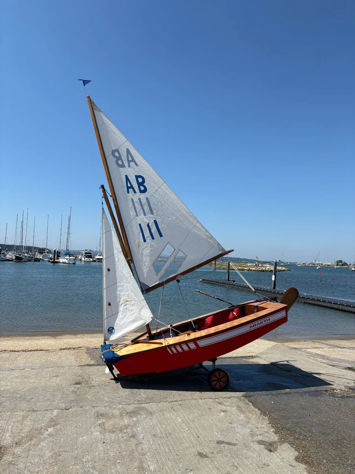 AB 111 Abarth rigged on her launching trolley on the slipway, red hull with a white stripe, varnished wooden decks and white sails, the harbour behind.