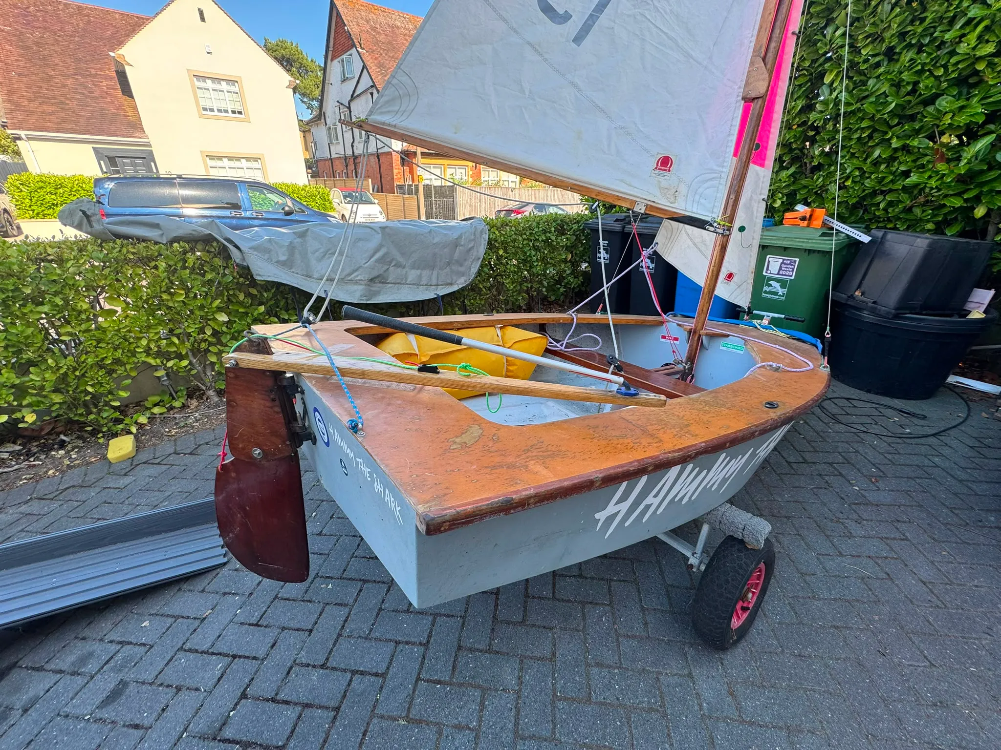 Looking forward over the stern quarter, the varnished wooden decks, a yellow buoyancy bag, the tiller and the dark wooden rudder.