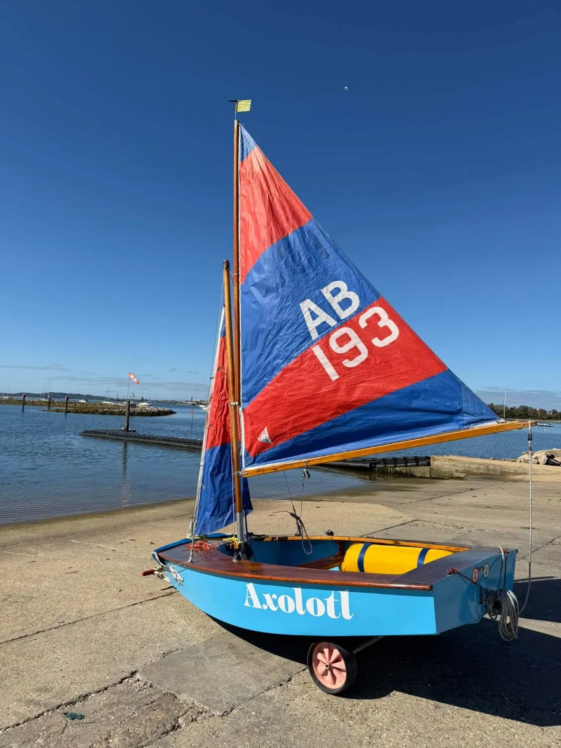 Axolotl, an AB with pale blue topsides, rigged on her trolley on the slipway with a red and blue mainsail and jib, Poole Harbour behind.