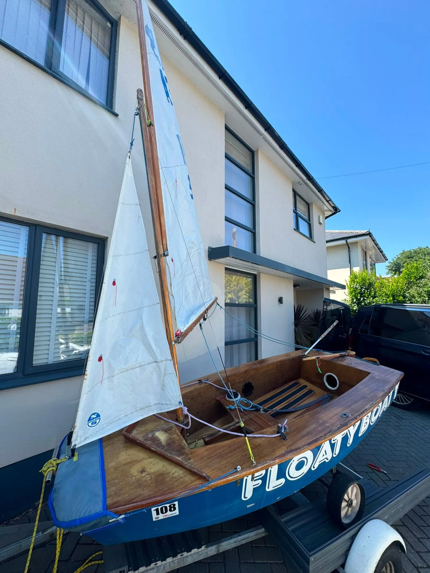 AB 311 Floaty Boaty rigged on her launching trolley, white North Sails main and jib hoisted on a varnished wooden mast, the name FLOATY BOATY in white on her dark blue topsides.