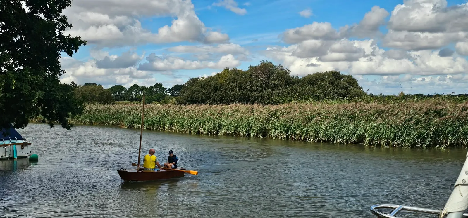 The same boat seen from astern, rowing away up the river between banks of tall reeds under a cloudy blue sky