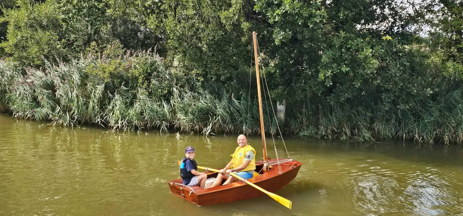 The varnished mahogany AB afloat on a reed-lined river, an adult in a yellow buoyancy aid rowing with pale wooden oars and a child sitting in the stern, her bare wooden mast stepped and stayed