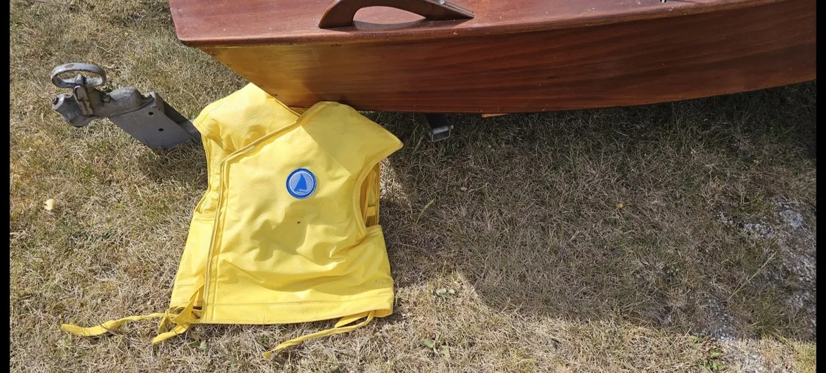 A yellow 1960s lifejacket, still with its ties, lying on the grass beside the bow of the mahogany hull and the trailer hitch