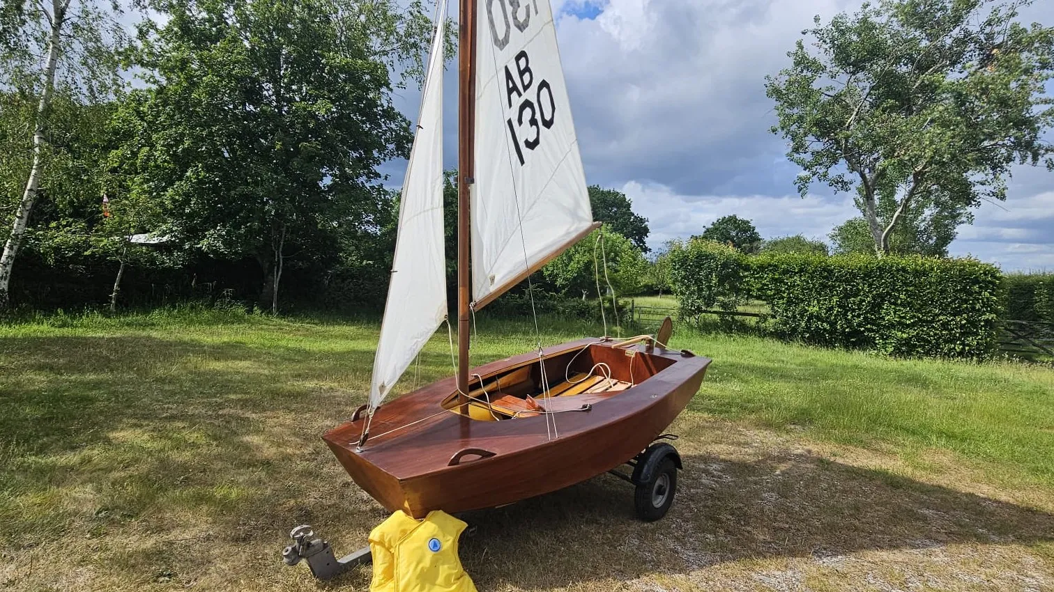 An original Poole AB rigged on her trailer on cut grass — varnished mahogany hull, Terylene sails, wooden spars, and a yellow lifejacket propped against the trailer hitch